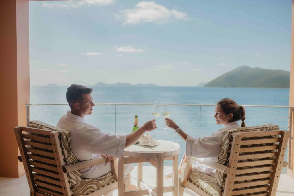 man and woman sitting in chairs on patio overlooking BVI waters while clicking champagne glasses